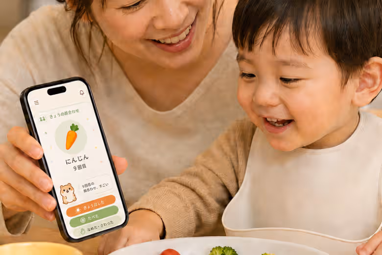 保護者がにんじん9回目の記録画面を見せ、服を着た幼児が笑顔を見せる温かい食卓の一場面